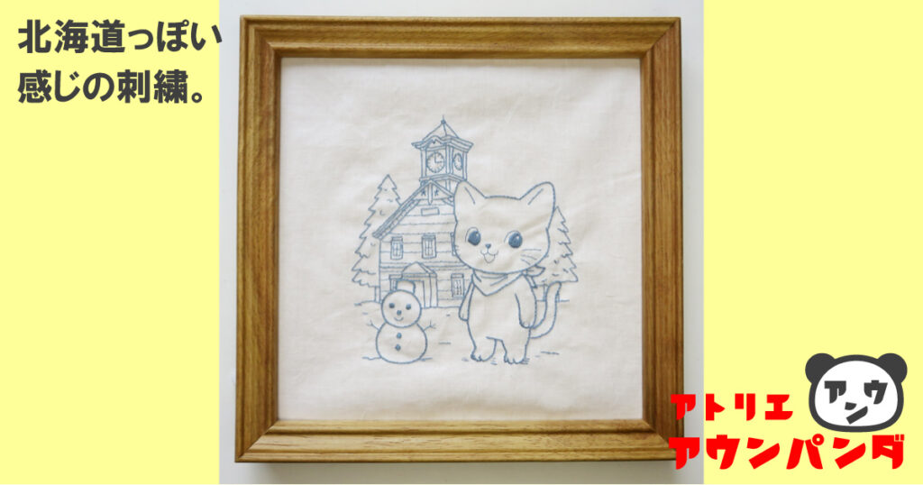 ブルー系の糸で一色刺繍した北海道モチーフの刺繍作品。猫と雪だるま、建物を描いた手刺繍